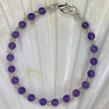 A beaded bracelet with purple and clear round beads and a silver clasp, displayed on a white seashell over a dark brown wooden surface.
