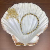 A beaded bracelet with small dark and light brown beads and a silver clasp, displayed inside a white scallop shell on a wooden surface.