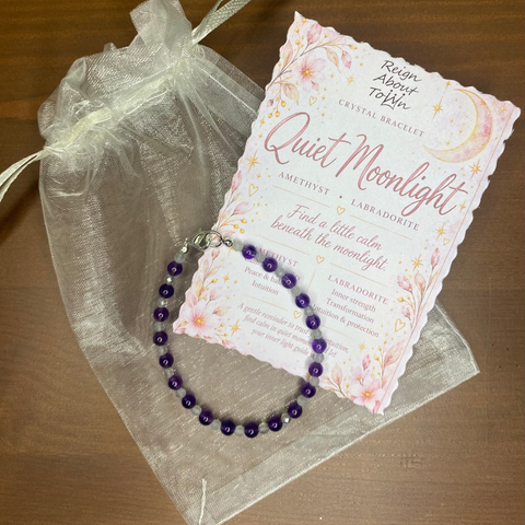 A purple crystal beaded bracelet with a silver clasp, presented on a sheer white organza gift bag next to a decorative card titled 'Quiet Moonlight' on a wooden tabletop.