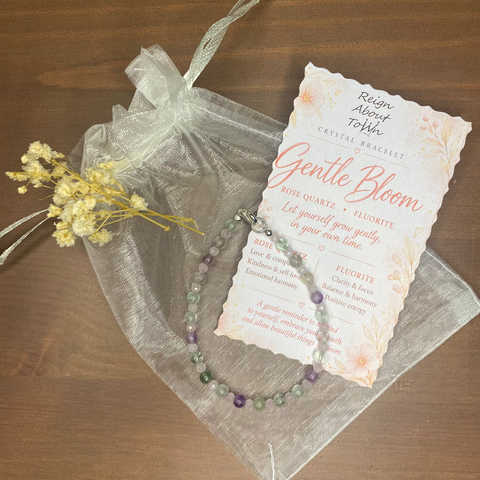 A beaded gemstone bracelet with purple and green beads, presented in a clear organza gift bag with a printed card that reads 'Gentle Bloom' on a wooden tabletop.